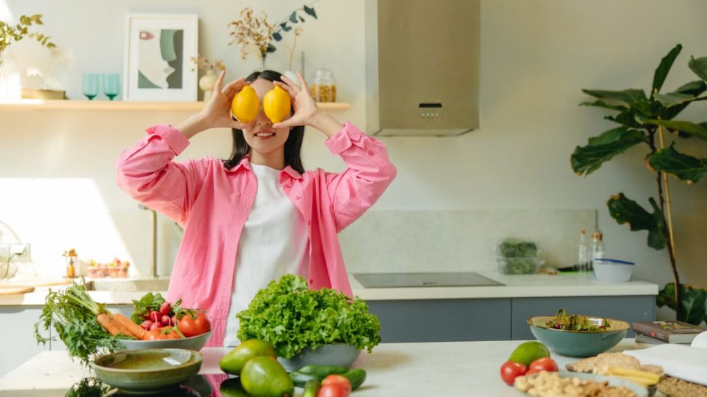 A woman playfully holding lemons over her eyes in a bright, lively kitchen setting filled with fresh vegetables.